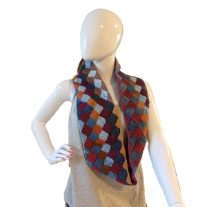 May include: A crochet infinity scarf featuring a multi-colored checkerboard pattern in shades of deep red, navy blue, light blue, and mustard yellow. The scarf is constructed with a textured, square-stitched design that forms a continuous loop.