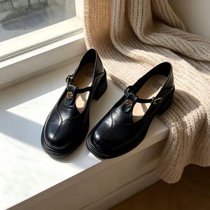 K&ouml;nnte beinhalten: Ein Paar schwarze Mary Jane-Schuhe mit runder Spitze und kleinem Blockabsatz. Die Schuhe haben einen Riemen &uuml;ber dem Fu&szlig; mit einer goldfarbenen Zierschnalle. Die Schuhe sind aus glattem, gl&auml;nzendem Material.