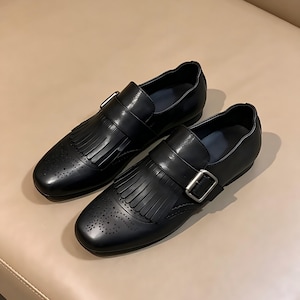 以下が含まれることがあります： バックル留めとフリンジのディテールが特徴の黒革靴のペア。靴は丸いつま先と小さな穴飾りのクラシックなデザインです。靴はニュートラルな表面に表示されています。