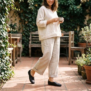 May include: A beige linen long-sleeve top with a V-neckline and matching beige linen wide-leg trousers with cuffed hems, paired with black woven leather flat shoes. The outfit features a relaxed fit and a natural, textured fabric appearance.
