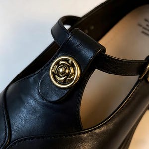 K&ouml;nnte beinhalten: Nahaufnahme eines schwarzen Mary Jane-Schuhs aus Leder mit einem goldfarbenen rosenf&ouml;rmigen Knopf am Riemen. Der Schuh hat ein T-Riemen-Design und einen Schnallenverschluss. Der Absatz ist im Hintergrund sichtbar.