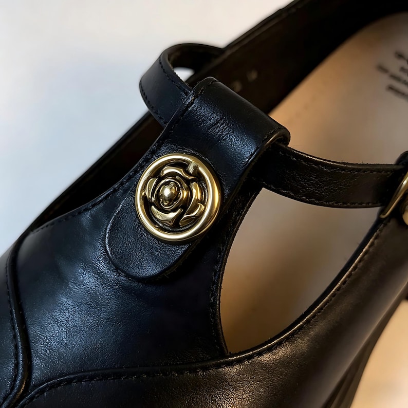 K&ouml;nnte beinhalten: Nahaufnahme eines schwarzen Mary Jane-Schuhs aus Leder mit einem goldfarbenen rosenf&ouml;rmigen Knopf am Riemen. Der Schuh hat ein T-Riemen-Design und einen Schnallenverschluss. Der Absatz ist im Hintergrund sichtbar.