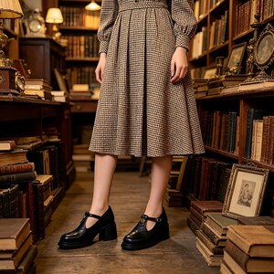 K&ouml;nnte beinhalten: Schwarze T-Steg-Schuhe aus Leder mit Blockabsatz. Die Schuhe haben einen Schnallenverschluss und eine runde Spitze. Die Schuhe werden mit einem braun-schwarz gemusterten Kleid in einer Bibliothek getragen.