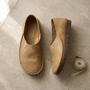 Könnte beinhalten: Ein Paar beigefarbene Schlupfschuhe aus Leder mit strukturierter Oberfläche und flachem Design. Die Schuhe liegen neben einem aufgerollten weißen Maßband, das die flexible Konstruktion und die abgerundete Zehenform der Schuhe zeigt.