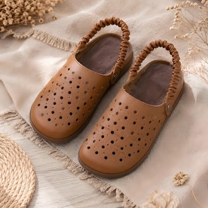 May include: A pair of tan slip-on clogs featuring a perforated upper with numerous circular cutouts for ventilation. Each shoe includes a gathered, elasticized heel strap attached with small metal rivets and a smooth, dark brown interior footbed.