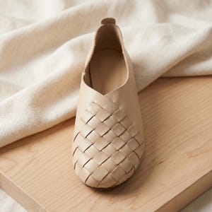 May include: A beige slip-on shoe featuring a woven leather upper with a grid pattern. The shoe has a rounded toe, a low-cut collar, and a smooth, unlined interior. The construction shows a seamless transition from the woven front to the solid heel.