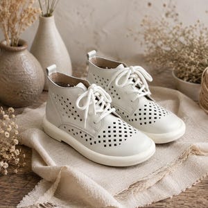 May include: A pair of off-white ankle boots featuring a laser-cut diamond pattern across the upper, white lace-up closures, a rounded toe, and a flat white rubber sole. The boots have a pull tab at the heel and a smooth, matte finish.