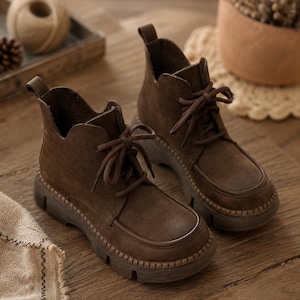 May include: A pair of dark brown ankle boots featuring a lace-up front, a rounded toe, and a thick, textured lug sole. The boots have a pull tab at the heel and a scalloped collar design, constructed with a matte finish material and matching laces.