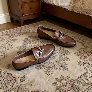 Può includere: Un paio di mocassini in pelle marrone con dettagli argentati. Le scarpe hanno un design classico con punta arrotondata e cinturino. Sono esposte su un tappeto fantasia. Calzature eleganti.