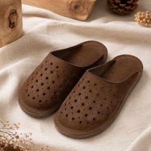 Könnte beinhalten: Ein Paar braune Slipper mit abgerundeter Zehenpartie und einem perforierten Muster auf dem Obermaterial. Die Hausschuhe haben eine durchgehende, flache Sohle und eine glatte, matte Oberfläche und sind als offene Schuhe für den Innenbereich gestaltet.