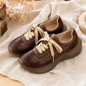 May include: A pair of dark brown sneakers featuring a perforated leather upper, suede-like toe and heel overlays, and thick, beige flat laces. The shoes have a chunky, molded brown rubber sole and a smooth, light-colored interior lining.