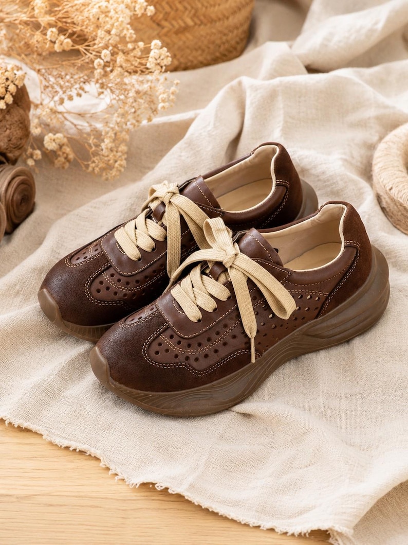 May include: A pair of dark brown sneakers featuring a perforated leather upper, suede-like toe and heel overlays, and thick, beige flat laces. The shoes have a chunky, molded brown rubber sole and a smooth, light-colored interior lining.