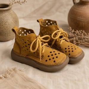 Puede incluir: Un par de botines color amarillo mostaza con un diseño perforado de aberturas circulares en la parte superior. Los botines tienen cordones amarillos a juego, punta redondeada y una suela de goma plana y gruesa en color marrón.