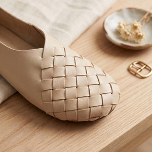 May include: A beige flat shoe featuring a woven leather upper with a rounded toe. The shoe has a smooth leather side panel and a structured design. The woven pattern covers the front portion of the shoe, creating a textured appearance.