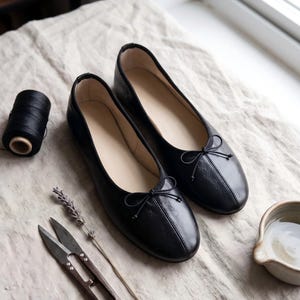 May include: A pair of black leather ballet flats with bow detailing. The shoes are presented on a textured, off-white surface, with a black thread spool, scissors, a lavender sprig, and a small ceramic bowl.