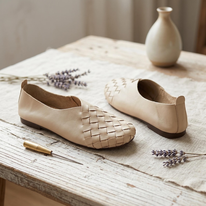 May include: A pair of beige slip-on shoes featuring a woven leather upper design, a rounded toe, and a low-profile dark brown sole. The shoes have a smooth heel tab and a cutout detail at the opening, showcasing a clean and simple construction.