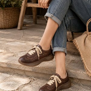 May include: A pair of dark brown leather sneakers featuring a perforated design on the toe box and sides, thick matching brown rubber soles, and wide cream-colored fabric laces tied in a bow across the top of the foot for a secure fit.