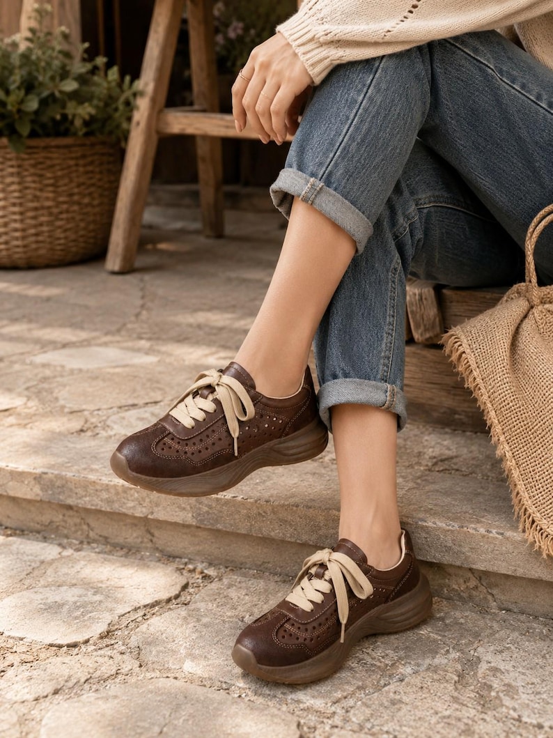 May include: A pair of dark brown leather sneakers featuring a perforated design on the toe box and sides, thick matching brown rubber soles, and wide cream-colored fabric laces tied in a bow across the top of the foot for a secure fit.