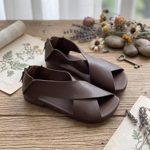 Peut inclure: Une paire de sandales plates marron foncé dotées d'une large bride croisée sur le dessus, d'un bout ouvert et d'une fermeture éclair verticale à l'arrière du talon. Les sandales présentent une finition lisse unie et une semelle basse.