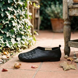 May include: A black slip-on shoe featuring a woven leather upper design on the front section, a smooth leather heel counter, a low-profile sole, and a small pull tab at the back of the collar, showcasing a clean and simple construction.