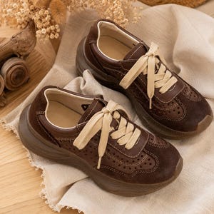 May include: A pair of dark brown sneakers featuring a thick platform sole, cream-colored laces, and perforated detailing along the sides and toe box. The shoes are constructed with a mix of smooth and suede-like textures and include visible stitching.