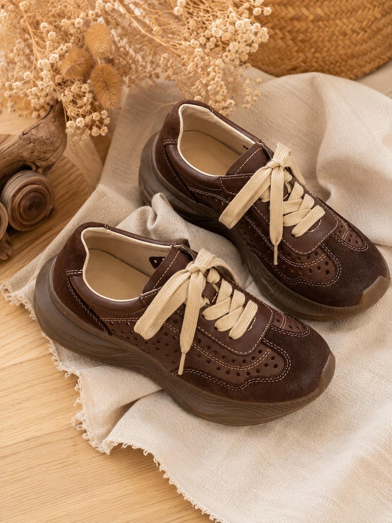 May include: A pair of dark brown sneakers featuring a thick platform sole, cream-colored laces, and perforated detailing along the sides and toe box. The shoes are constructed with a mix of smooth and suede-like textures and include visible stitching.