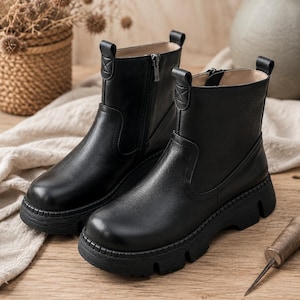 May include: A pair of black ankle boots featuring a smooth leather-like finish, a rounded toe, a side zip closure, and a thick, lugged rubber sole. Each boot includes a pull tab at the heel and stitched detailing on the upper shaft.