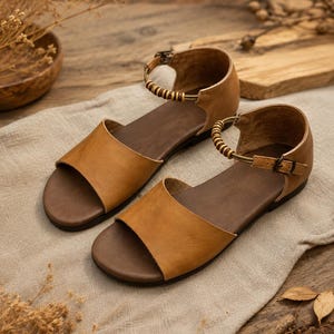 May include: A pair of tan leather sandals featuring an open-toe design, a dark brown footbed, and an ankle strap secured with a small metal buckle. The ankle strap is accented with a series of small, cylindrical wooden beads along the side.