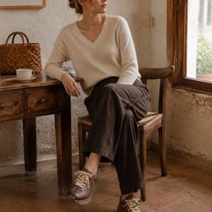 May include: A cream-colored long-sleeve V-neck sweater with a ribbed texture, paired with dark brown wide-leg trousers and brown leather sneakers featuring decorative perforations and thick cream-colored laces.