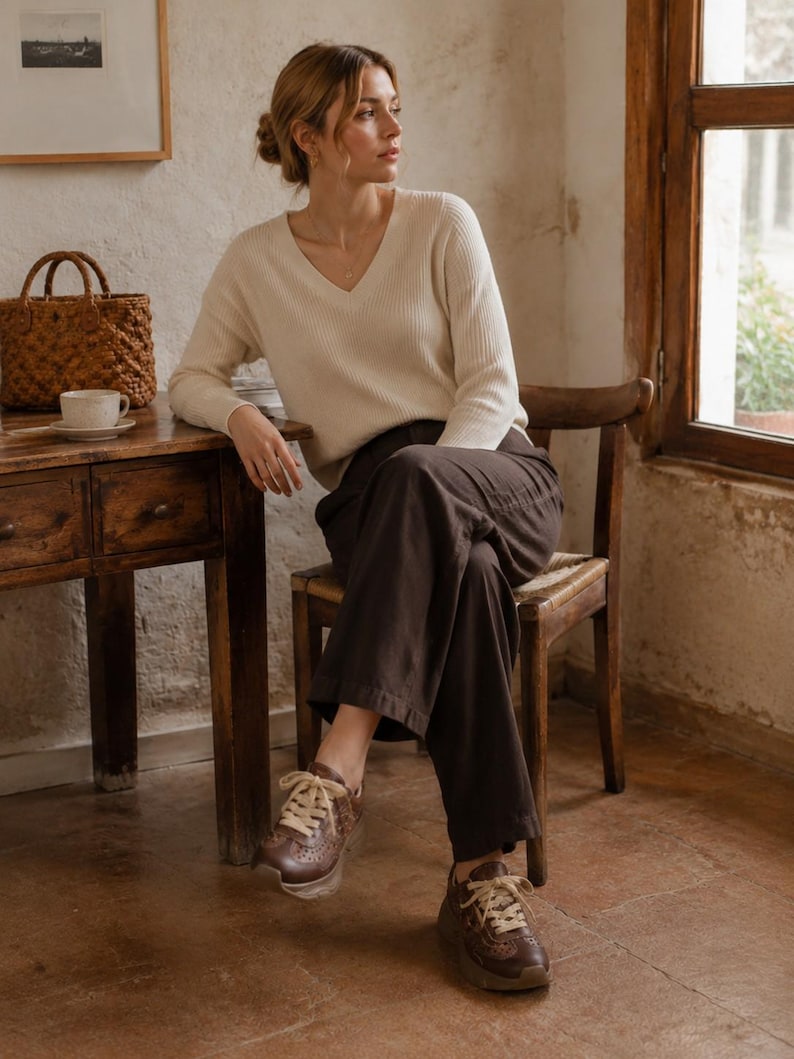 May include: A cream-colored long-sleeve V-neck sweater with a ribbed texture, paired with dark brown wide-leg trousers and brown leather sneakers featuring decorative perforations and thick cream-colored laces.