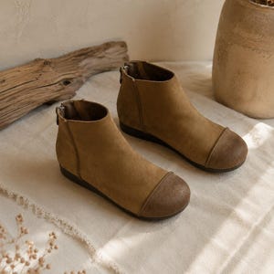 May include: A pair of tan suede ankle boots featuring a rounded toe cap with a darker, textured finish. The boots have a flat sole and a vertical zipper closure at the back of the heel, providing a clean and simple silhouette for everyday wear.