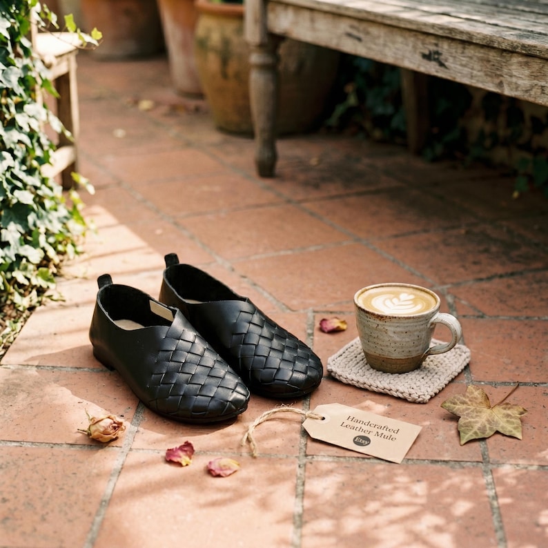 May include: A pair of black leather mules with a woven upper design, a ceramic mug containing a latte with heart-shaped foam art on a beige coaster, and a tan paper tag labeled Handcrafted Leather Mule with the Etsy logo printed on it.