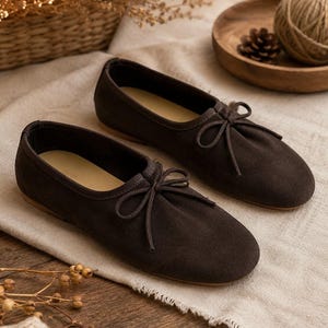 Peut inclure: Une paire de ballerines en daim marron foncé avec un bout arrondi, une encolure basse et un fin nœud décoratif en cordon noué au centre de l'empeigne. Les chaussures présentent une construction plate simple avec une doublure intérieure lisse.