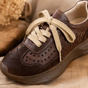 May include: A dark brown sneaker featuring a perforated leather upper, a suede toe cap, and thick beige laces. The shoe includes a chunky, molded brown rubber sole with a wavy texture and a light-colored interior lining.