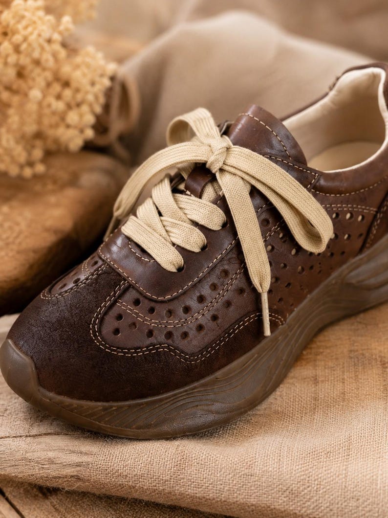 May include: A dark brown sneaker featuring a perforated leather upper, a suede toe cap, and thick beige laces. The shoe includes a chunky, molded brown rubber sole with a wavy texture and a light-colored interior lining.
