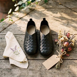 May include: A pair of black woven leather slip-on shoes with a rounded toe and a low-cut opening. The shoes are positioned next to a cream-coloured cloth featuring embroidered floral sprigs and a small bouquet of dried flowers with a blank tag.