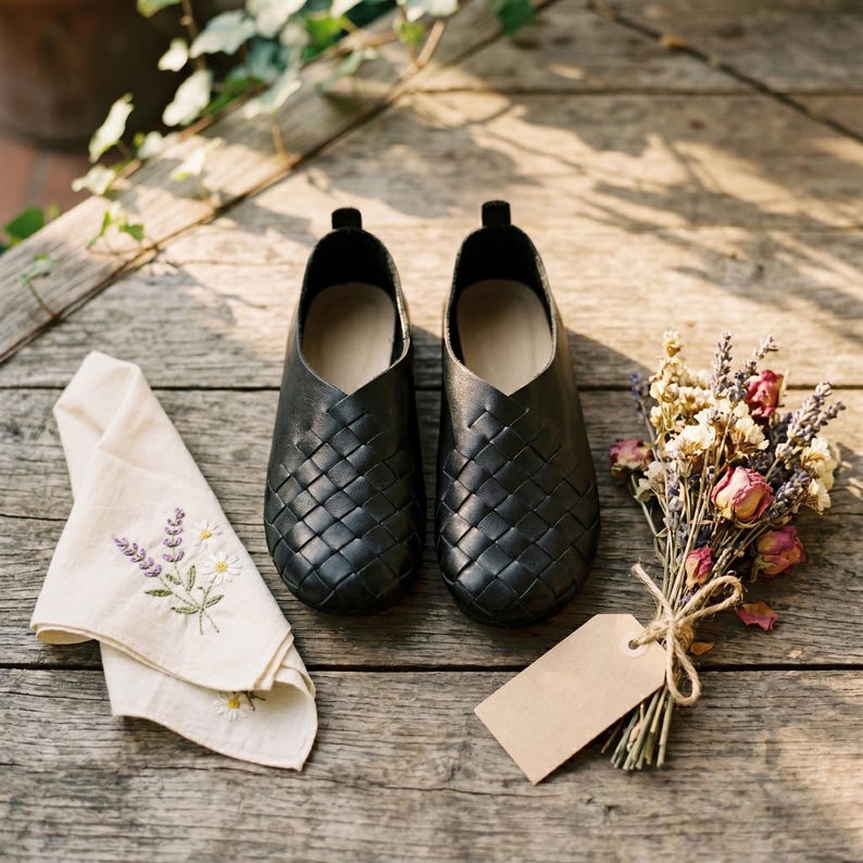May include: A pair of black woven leather slip-on shoes with a rounded toe and a low-cut opening. The shoes are positioned next to a cream-colored cloth featuring embroidered floral sprigs and a small bouquet of dried flowers with a blank tag.