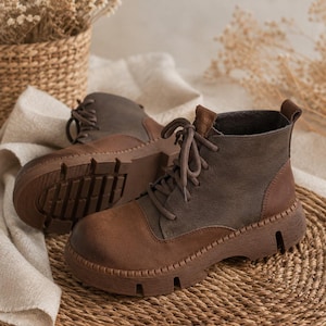 May include: A pair of ankle boots featuring a two-tone design with dark brown leather toe caps and heel counters, contrasted by olive green leather side panels. The boots include matching brown laces, a pull tab at the heel, and thick lug soles.