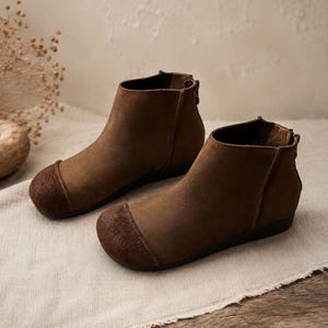 Puede incluir: Un par de botines marrones con parte superior de cuero liso y puntera de ante texturizado en un tono más oscuro. Los botines tienen un perfil bajo, una suela plana negra y un cierre de cremallera vertical situado en la parte posterior del talón.