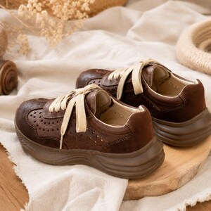 May include: A pair of dark brown sneakers featuring a perforated leather upper, suede heel panels, thick matching brown rubber soles, and cream-colored flat laces. The shoes have a low-top design with visible stitching along the side panels.