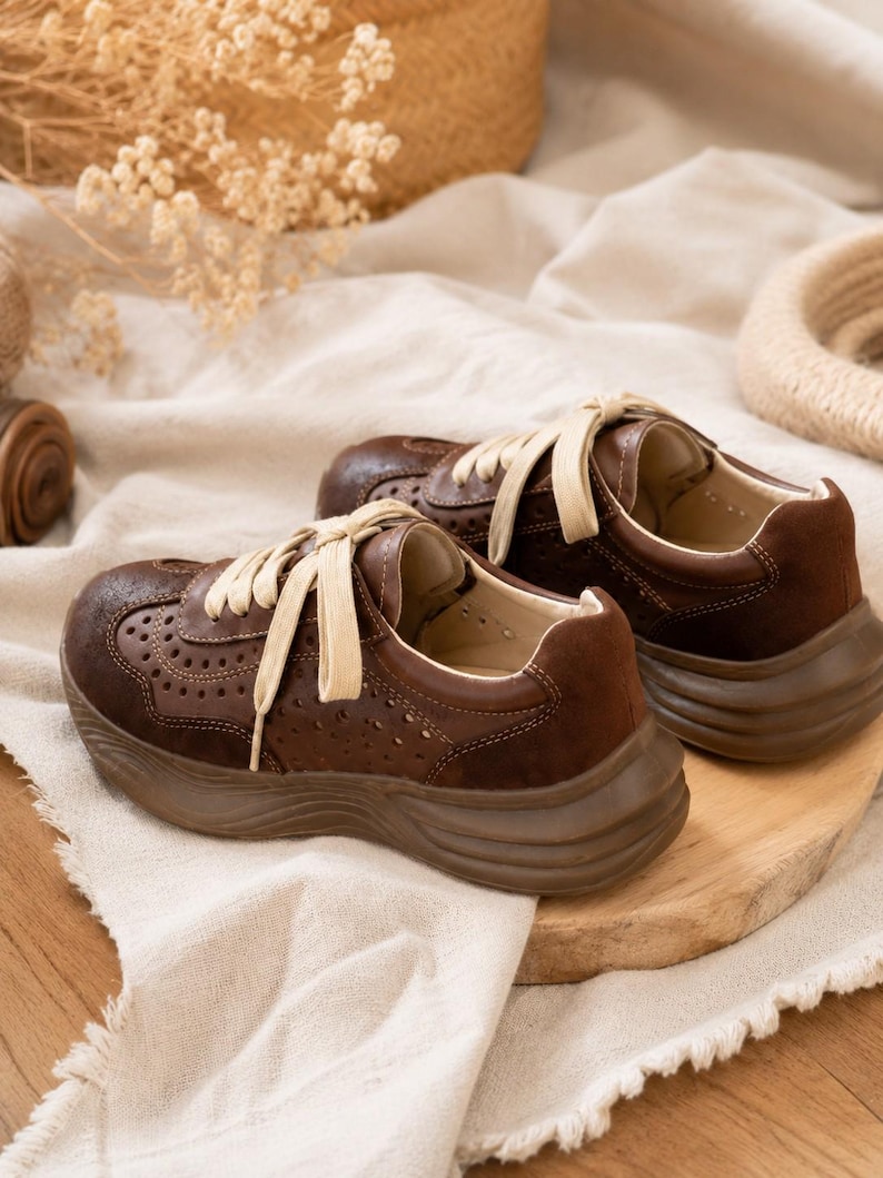 May include: A pair of dark brown sneakers featuring a perforated leather upper, suede heel panels, thick matching brown rubber soles, and cream-colored flat laces. The shoes have a low-top design with visible stitching along the side panels.