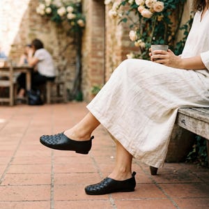 May include: A pair of black leather slip-on shoes featuring a woven texture on the front toe area and a smooth finish on the heel. The shoes have a low profile, a rounded toe, and a flat sole, providing a simple and functional footwear design.