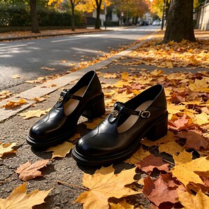 K&ouml;nnte beinhalten: Schwarze Mary Jane-Schuhe aus Leder mit einem klobigen Absatz. Die Schuhe haben ein T-Riemen-Design mit einer goldfarbenen Schnalle und einem Emblem. Die Schuhe liegen auf einer Oberfl&auml;che mit Herbstbl&auml;ttern.