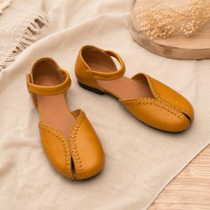 May include: A pair of mustard yellow flat shoes featuring a rounded toe, a cutout design along the center, and a decorative stitched seam detail. The shoes include a thin ankle strap with a side closure and a low, flat heel for a simple silhouette.