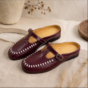 May include: A pair of dark burgundy leather mules featuring a T-strap design with a silver-toned metal buckle. The rounded toe area is accented with prominent white whipstitching along the edge, and the interior is lined with a light tan material.
