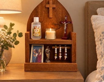 Puede incluir: Un altar doméstico de madera con una parte superior en arco apuntado y una cruz central. El estante superior contiene una botella de agua bendita de Knock, una vela encendida en un frasco de vidrio y un crucifijo. El estante inferior muestra una tarjeta religiosa, una figura y tres rosarios.