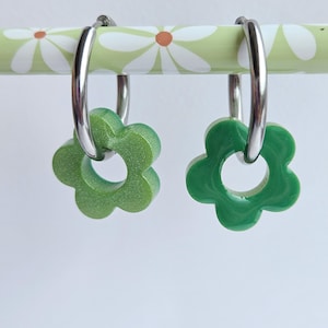 Peut inclure: Une paire de boucles d'oreilles créoles argentées, chacune dotée d'une breloque en forme de fleur. L'une est d'un vert clair scintillant, l'autre d'un vert foncé brillant et uni. Les boucles sont suspendues à une tige vert clair à motifs de marguerites.