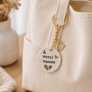 Può includere: Borsa tote color crema con portachiavi dorato e ciondolo rotondo. Il ciondolo riporta la scritta "Merci Maman" in nero, con dettagli a cuore e stampa leopardata. La borsa ha una tasca frontale e un manico.