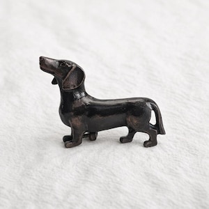 May include: A bronze-colored dachshund figurine. The dog is posed with its head raised, ears perked, and tail slightly curved. The figurine has a glossy finish and is set against a white background.