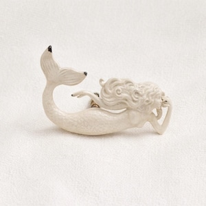 May include: Ivory-colored mermaid figurine with a flowing tail and long, curly hair. The mermaid is in a reclining pose, with one arm resting on her head. The tail and the tips of the fins are painted black. This decorative item is suitable for home decor.